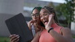 © Marco - Two young women speaking in video with family holding tablet outdoors. African American adult girl waving hello using modern device communication. Girlfriends with box braids hairstyle