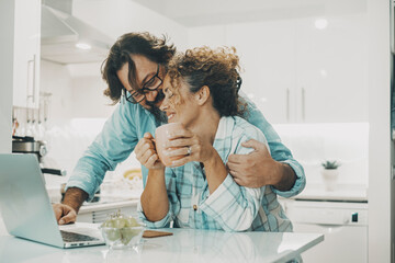 Two people man and woman couple at home enjoying indoor leisure activity in the kitchen using laptop computer to  surf the web. Indoor real lifestyle for young mature female male in love relationship