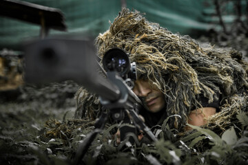 Man in a ghillie suit hidden in the forrest, aiming through a scope on a rifle. May both represent a sniper or a hunter, special forces soldier assault rifle with silencer. Sniper in the forest.