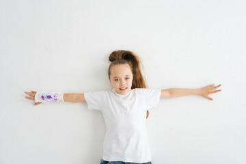 Wall Mural - Top view of cheerful cute little girl with broken hand wrapped in plaster bandage with colorful draw lying on white floor in studio looking at camera. Concept of child insurance and healthcare