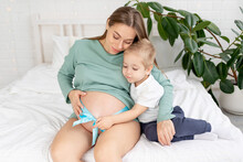 A Pregnant Woman And A Baby Son Touches A Big Belly Tied With A Blue Ribbon At Home And Cuddle On The Bed, The Concept Of Pregnancy And Waiting For The Birth Of A Baby And A Second Child In The Family