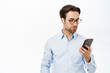 Handsome businessman in glasses, using his mobile phone, working on smartphone app, standing in blue collar shirt over white background