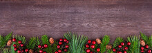 Winter Bottom Border With Evergreen Branches, Red Berries And Pine Cones. Top View On A Dark Wood Banner Background With Copy Space.