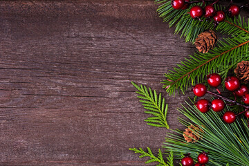  Christmas side border with evergreen branches, red berries and pine cones. Overhead view on a dark wood background with copy space.