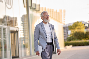 Happy cool bearded stylish old mature professional business man, smiling gray haired senior older businessman wearing suit holding tablet walking going outdoors in big city business district downtown.