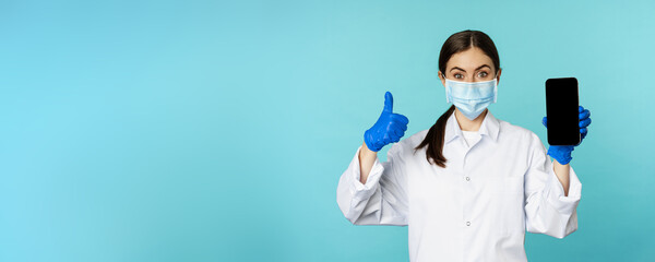 Portrait of doctor in medical face mask and gloves, showing mobile phone app, smartphone screen and thumb up, recommending online checkup website, standing over blue background