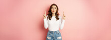 Portrait Of Surprised Caucasian Woman, Stylish Girl Pointing Fingers Up And Looking Impressed, Showing Store Logo, Discount Advertisement, Pink Background