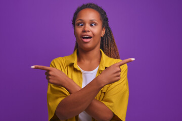 Wall Mural - Young funny African American woman dressed in casual style looks and shows with hands different sides of at same time cannot make choice or fool around, stands on plain purple background.