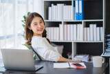 Fototapeta  - Beautiful Asian businesswoman working on a desk with a laptop with a smiling face while looking at the document received in the office.