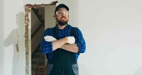 Wall Mural - Smiling man in baseball cap wearing protective suit and plaid shirt has goggles on to protect eyes hands crossed over chest stands in middle of renovated house.