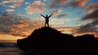 © Valua Vitaly - Man stands on a rock against the ocean during summer sunset. Human with raised hands looks to sun over horizon in morning while sunrise. Man feels happy and freedom. Silhouette of a man against  sky
