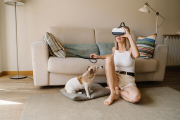 blond woman testing wearing virtual reality simulator headset home small dog Jack Russell terrier sitting next to her watching confused. escape from reality theme. Light home living room with sofa