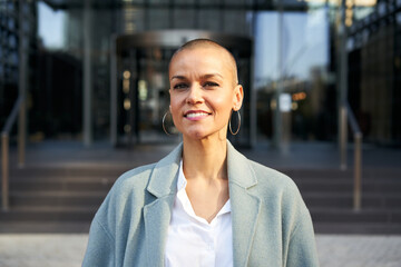 Cheerful middle-aged empowered woman with shaved hair looking at camera. Business people outdoors on background with buildings. corporate. High quality photo