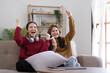 © wichayada - Two Young womanWatching TV Shaking Fists In Joy Celebrating Victory Of Favorite Sport Team Sitting On Couch In Living Room At Home. Weekend Leisure, Television Show And Entertainment Concept