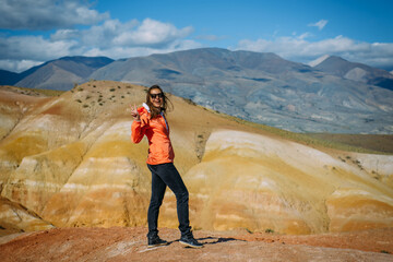 Happy young woman standing on a rock with beautiful view. Cheerful female traveller gesturing at camera on stunning mountains background. Freedom lifestyle, wanderlust theme.