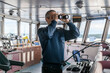 Deck officer with binoculars on navigational bridge. Seaman on board of vessel. Commercial shipping. Cargo ship.