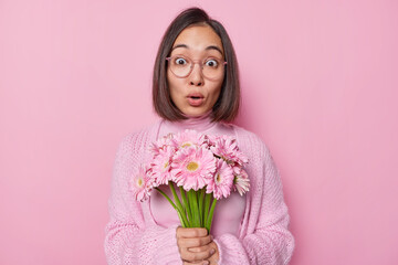 Wall Mural - Shocked Asian woman with dark hair holds breath cannot believe own eyes receives beautiful bouquet of gerberas wears optical eyeglasses and casual knitted jumper isolated over pink background