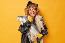Horizontal Shot Of Overjoyed Curly Haired Female Angler Carries Big Trophy Caught Salmon Laughs Happily Wears Black Leather Raincoat And Hat Carries Fishing Net Isolated Over Yellow Background