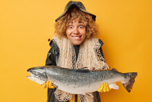 Positive Fisherwoman With Curly Hair Bites Lips Holds Huge Caught Salmon Carries Fishnet Around Neck Dressed In Black Leather Raincoat Isolated Over Vivid Yellow Background. Fishing And Recreation