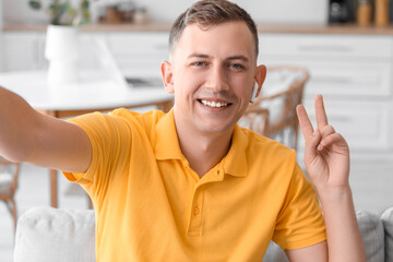 Sticker - Young man showing victory gesture while taking selfie on sofa at home