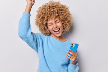 Sweet success. Joyful curly haired young woman makes victory gesture holds mobile phone glad to pass online test successfully receives good news wears blue jumper isolated over grey background