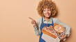 © WHstudio Leushin N - Happy female baker wears shirt and denim sarafan works in bakery store holds freshly baked bread on linen towel bites lips points index finger aside demonstrates blank space for your advertisement