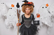 Funny curly haired female wizard with spider on face crosses eyes makes grimace holds broom and cauldron involved in witchcraft wears black hat dress poses around spooky ghosts. Halloween is coming