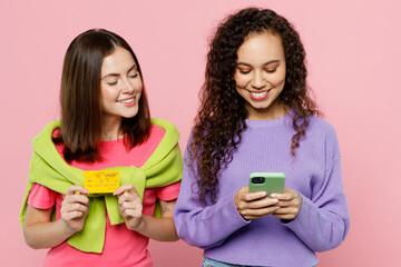 Wall Mural - Young two friends women wearing green purple shirts looking camera together using mobile cell phone hold credit bank card shopping online order delivery isolated on pastel plain light pink background.