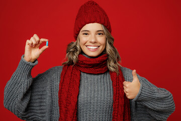 Wall Mural - Young smiling fun woman wear grey sweater scarf hat hold in hand blue pill thumb up isolated on plain red background studio portrait. Healthy lifestyle ill sick disease treatment cold season concept.