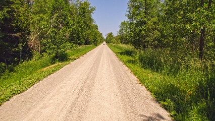 Aerial of white family SUV hatchback moving through green rural countryside landscape by gravel road. Green flat American forests on both sides of the road. Travelling and vacation.