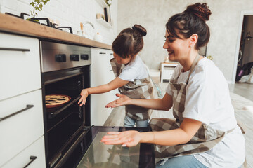 Wall Mural - A little girl and her mom put a baking dish with a pie in the oven in the kitchen. mom and child cook a pie together in the kitchen.