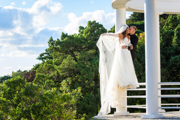 Wall Mural - The newlyweds happily embrace in a beautiful gazebo with columns against the backdrop of foliage and sky