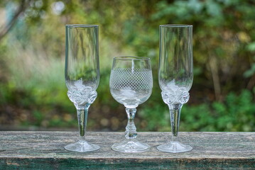 a row of white three glass crystal goblets on a gray wooden table outdoors on a green background