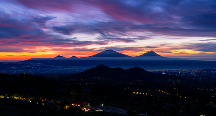 Wall Mural - Beautiful colorful epic sunrise sky with mountain range and beautiful city lights, Magelang City and Merapi, Merbabu, Andong, Telomoyo mountain looking out from Sumbing Mountain - Mangli Village