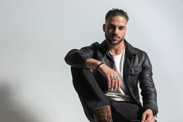 Wall Mural - stylish young man wearing black leather jacket posing with elbow on knee