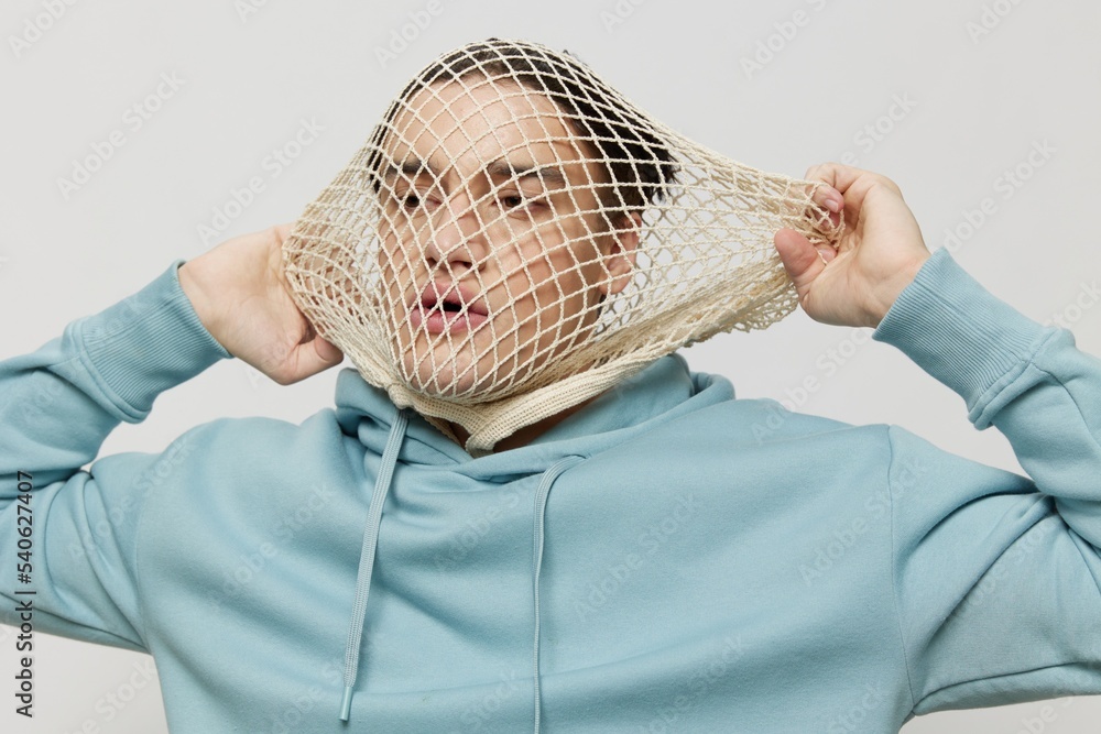 tortured, suffering from problems, a man with a mesh on his face stands in a light blue hoodie on a light background and pulls it aside trying to take it off