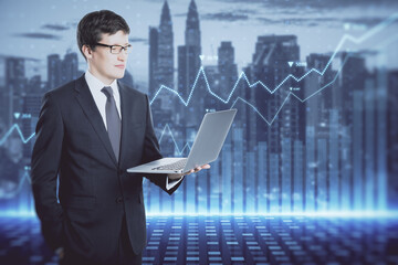 Poster - Attractive young european businessman with laptop and abstract glowing blue business graph hologram on blurry city background. Economy, finance and digital money concept. Double exposure.