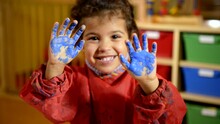Kids, School And Education With Happy Children Having Fun And Painting With Hands In Kindergarten. Female Child Hand Painting On Paper. Portrait Of Little Girl Smiling At Camera At Preschool