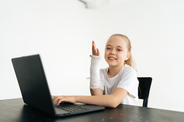 Wall Mural - Cheerful blonde little girl raising broken hand wrapped in white plaster bandage sitting at table with laptop smiling looking at camera in light room. Concept of child insurance and healthcare.