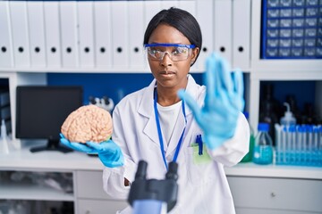 Poster - African young woman working at scientist laboratory holding brain with open hand doing stop sign with serious and confident expression, defense gesture