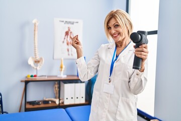 Sticker - Middle age blonde woman holding therapy massage gun at physiotherapy center smiling happy pointing with hand and finger to the side