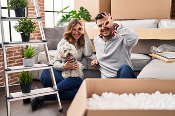 Poster - Young hispanic couple sitting on the floor at new home with log smiling happy doing ok sign with hand on eye looking through fingers