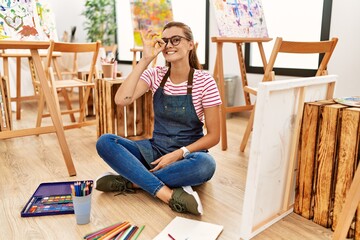 Poster - Young brunette woman at art studio sitting on the floor doing ok gesture with hand smiling, eye looking through fingers with happy face.