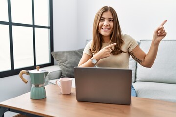 Canvas Print - Young brunette woman using laptop at home drinking a cup of coffee smiling and looking at the camera pointing with two hands and fingers to the side.