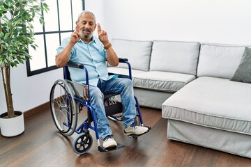 Canvas Print - Handsome senior man sitting on wheelchair at the living room gesturing finger crossed smiling with hope and eyes closed. luck and superstitious concept.