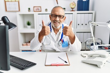 Wall Mural - Mature doctor man at the clinic success sign doing positive gesture with hand, thumbs up smiling and happy. cheerful expression and winner gesture.