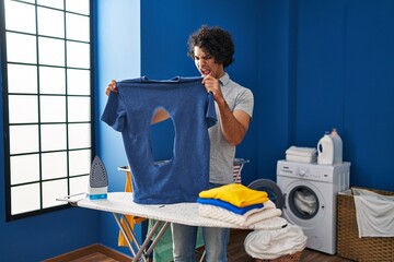 Wall Mural - Hispanic man with curly hair ironing holding burned iron shirt at laundry room angry and mad screaming frustrated and furious, shouting with anger. rage and aggressive concept.