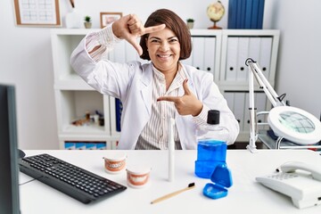 Canvas Print - Middle age hispanic dentist woman working at medical clinic smiling making frame with hands and fingers with happy face. creativity and photography concept.