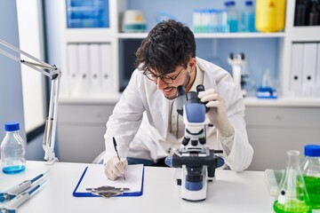 Wall Mural - Young hispanic man scientist writing on document using microscope at laboratory