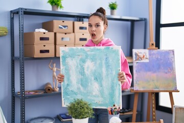 Poster - Young hispanic girl holding canvas in shock face, looking skeptical and sarcastic, surprised with open mouth